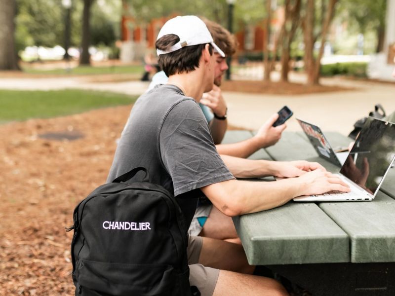 Chandelier Student Living Apartments Two people sit at a picnic table outside, one using a laptop, with a black “Chandelier” backpack in the foreground.